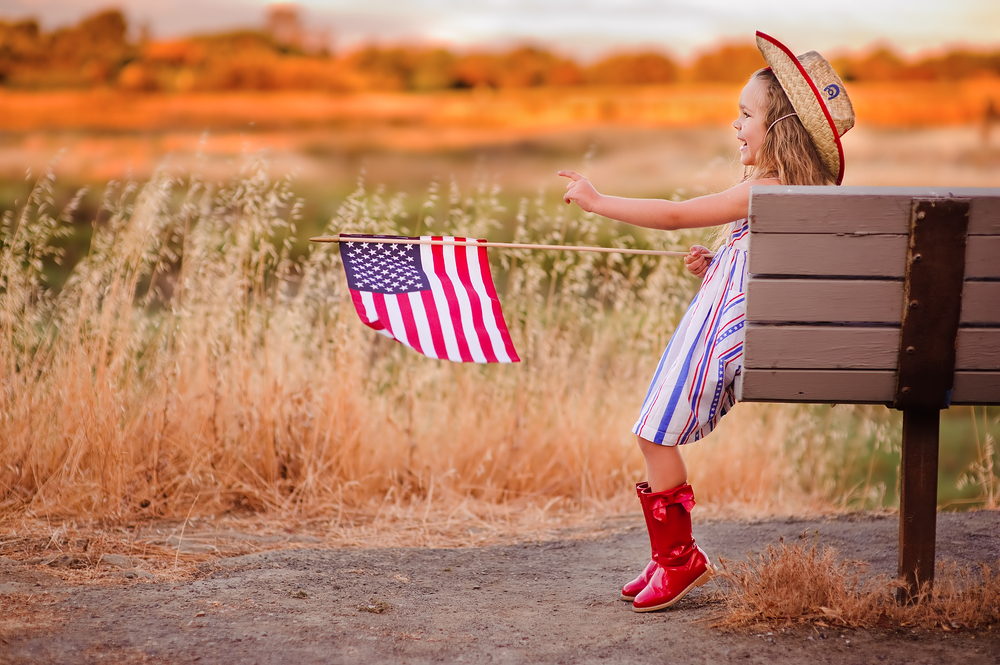 A young girl in red boots sitting on a bench and holding an American flag