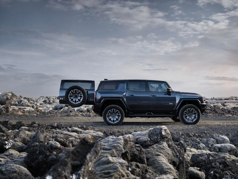 Passenger side view of a slate blue 2025 GMC Hummer EV SUV with the trunk open and parked on rocky terrain at dusk